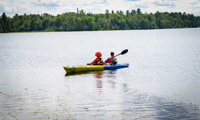 Two people paddling in a tandem kayak on a lake, surrounded by wilderness on all sides