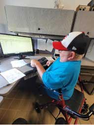Physically disabled man in blue shirt and baseball cap sits on a walker and types at a computer station