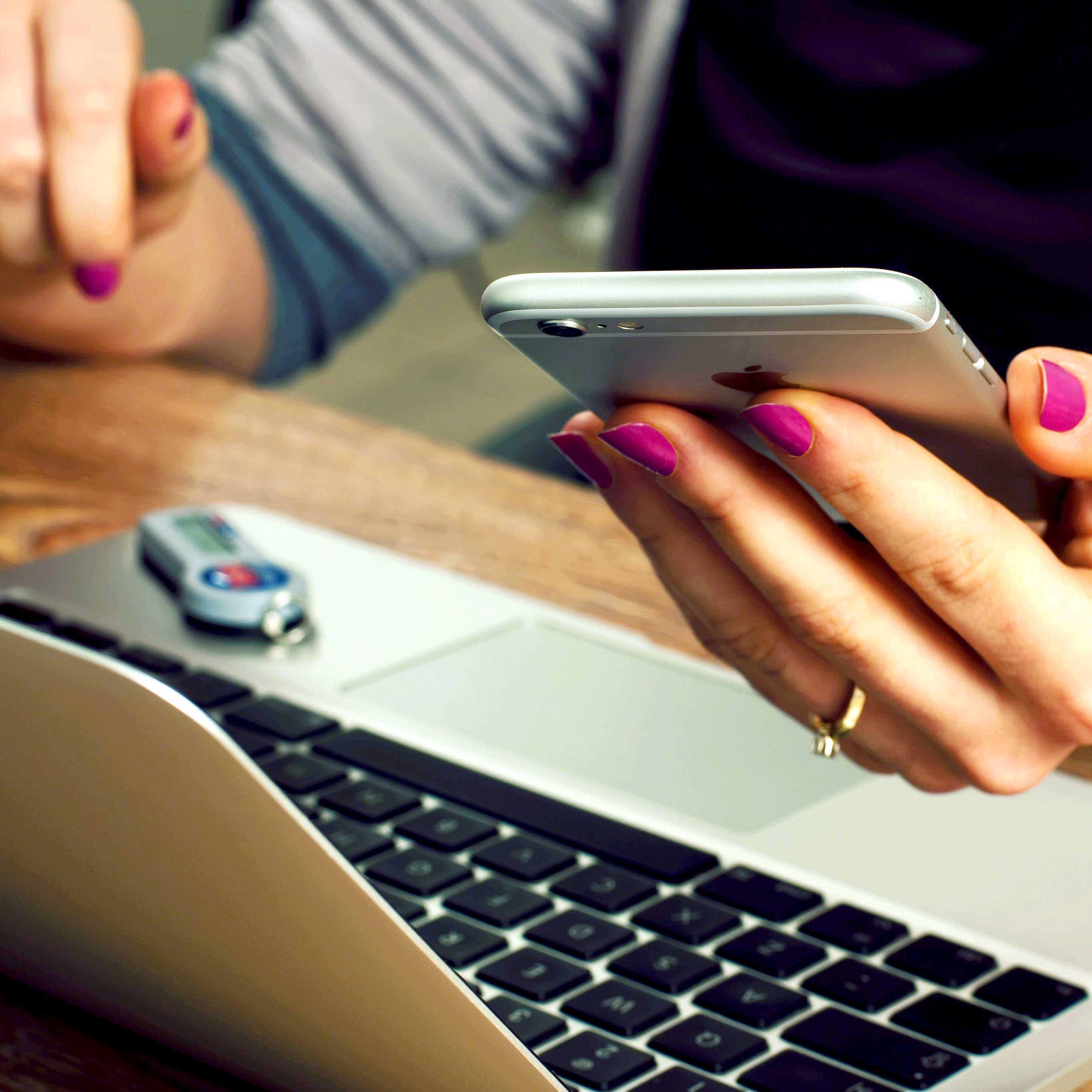 Tight image of a woman holding a mobile phone in one hand, the other hand poised over a laptop keyboard