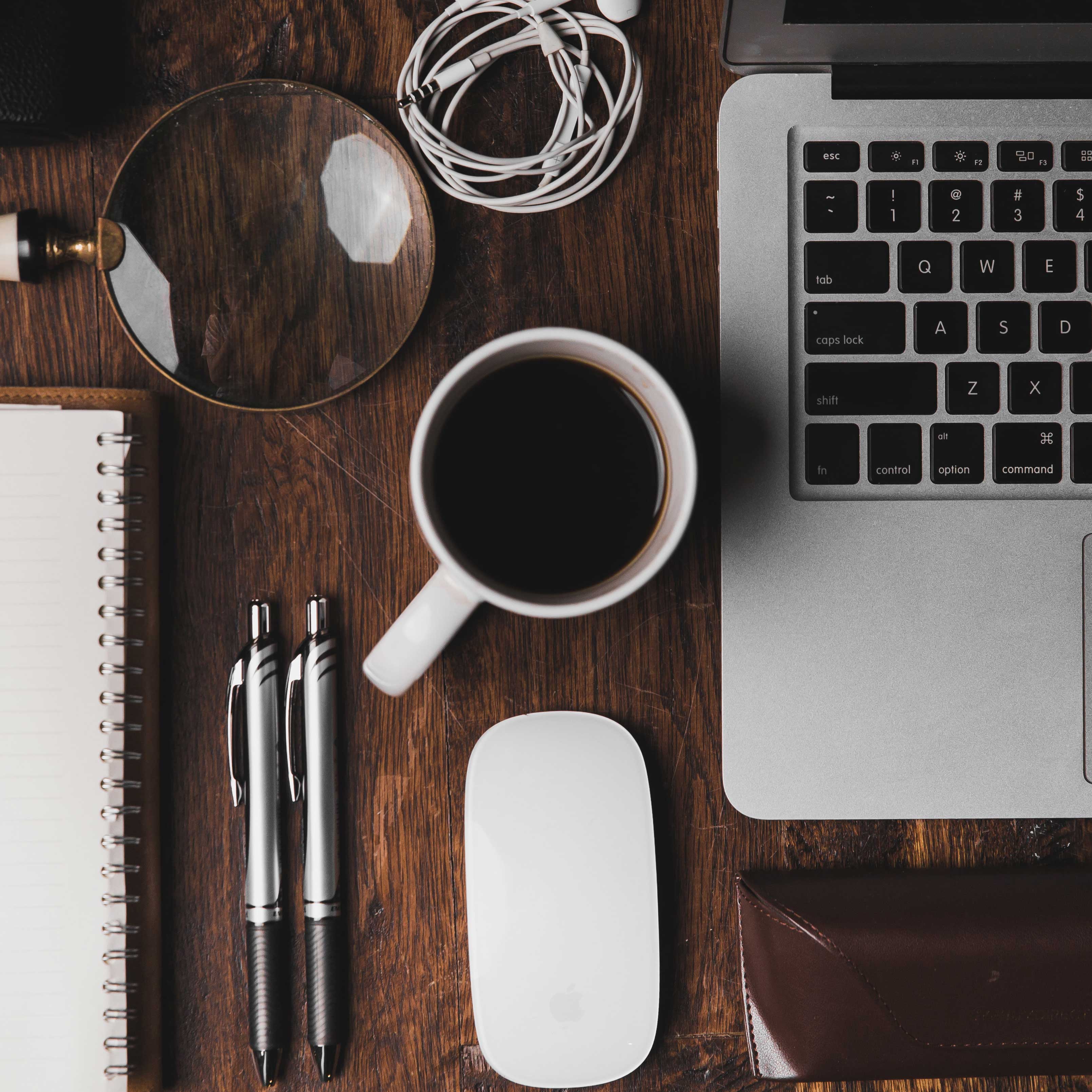 Overhead view of a desktop, with a computer, mouse, pens, pad of paper, headphones and a cup of coffee arranged on it