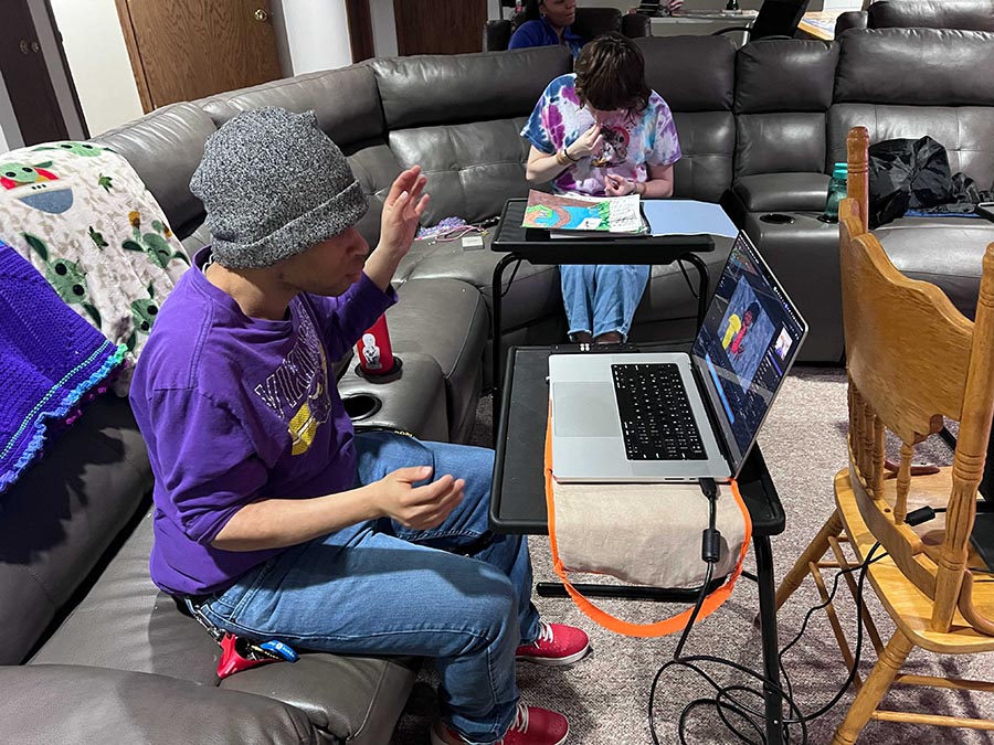 Two men, one in a Vikings sweatshirt and gray knit cap, and the other wearing a tie-dyed T-shirt, sit on a sectional sofa and work on separate computer work stations