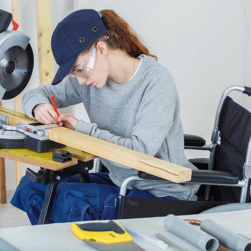 Woman in baseball cap and safety glasses and using a wheelchair works over a drafting table in a workshop. In the foreground are handtools on a workbench. A miter saw is visible in the background